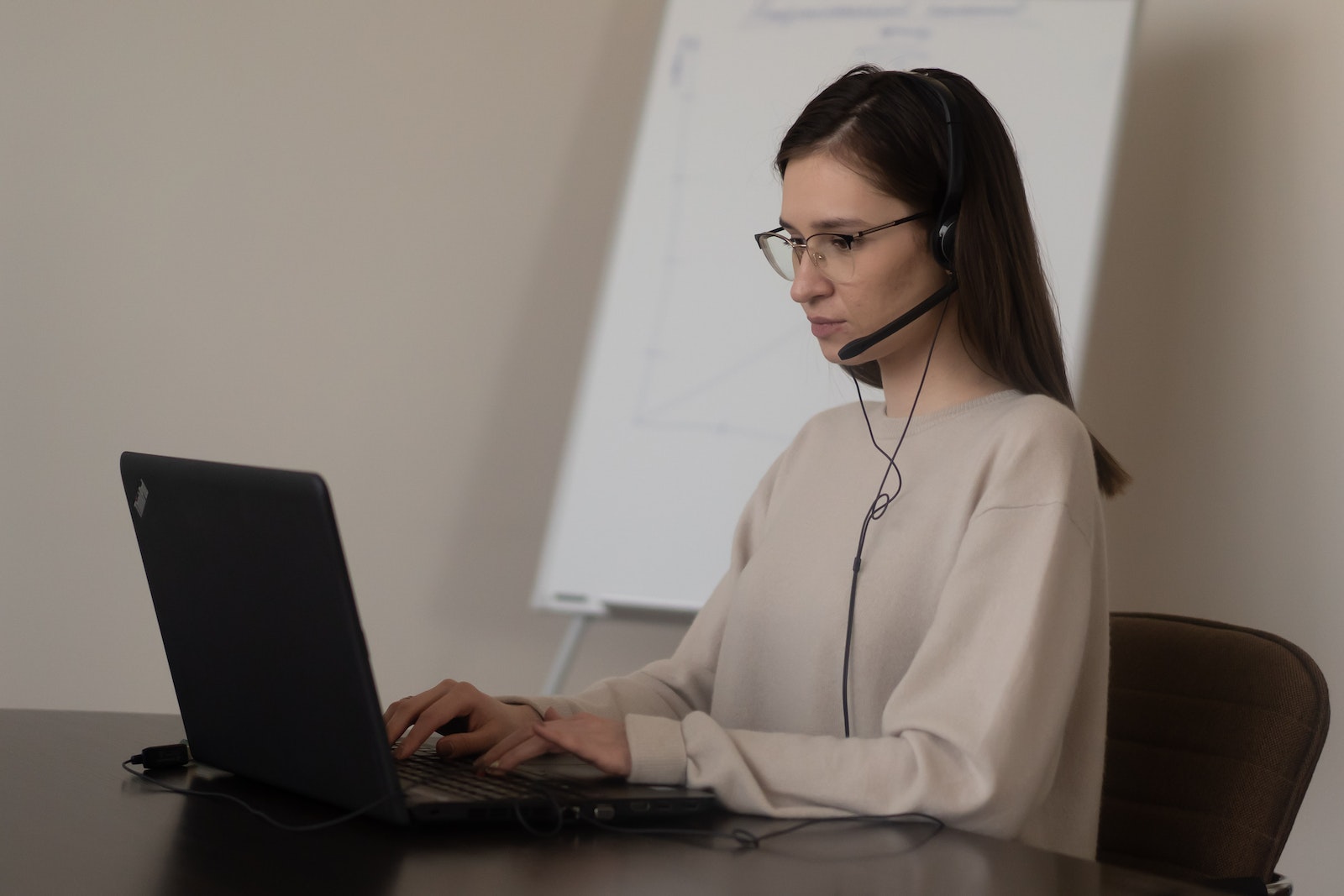 Jeune femme sérieuse en tenue décontractée avec des lunettes, assise sur une chaise à une table et travaillant sur un ordinateur portable avec des écouteurs et un microphone près d'un tableau blanc sur un lieu de travail lumineux.