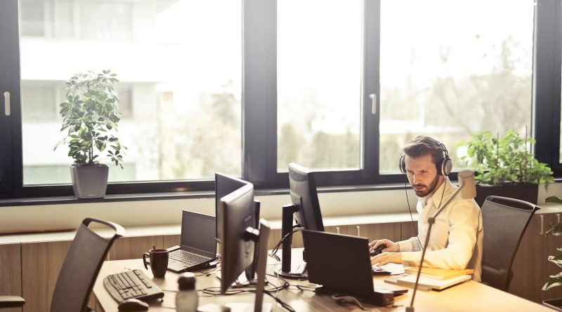 Un homme d'affaires est assis à un bureau, utilisant plusieurs ordinateurs et un casque, dans un bureau moderne bien éclairé.